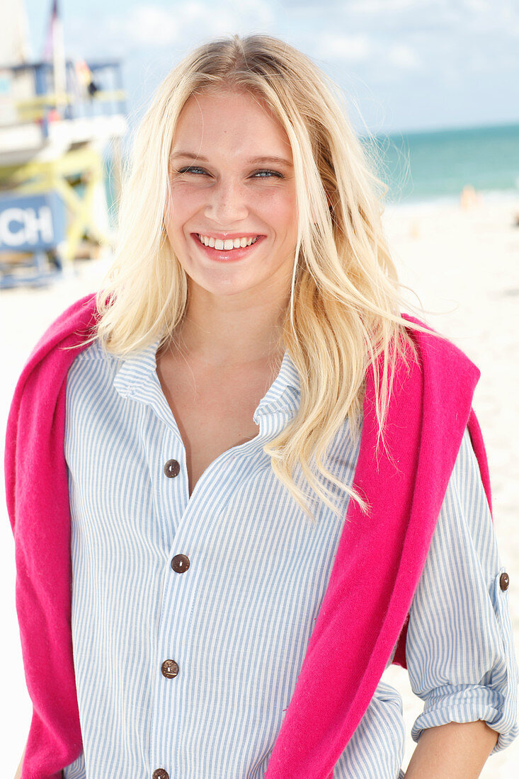 Junge blonde Frau in gestreiftem Hemd und mit pinkfarbenem Pulli am Strand