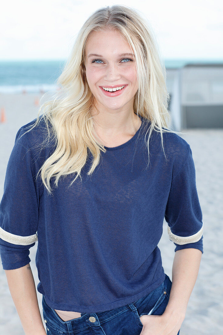 A young blonde woman on a beach wearing a long-sleeved blue shirt