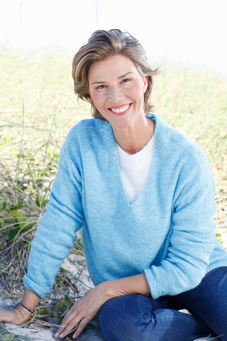 A mature woman with short blonde hair in the countryside wearing blue jeans and a blue jumper over a white shirt