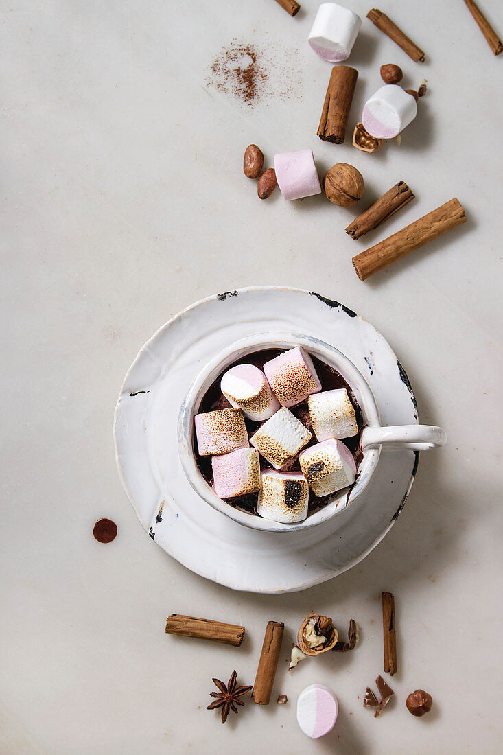 Ceramic cup of hot chocolate with marshmallow s'mores with ingredients above over white marble table