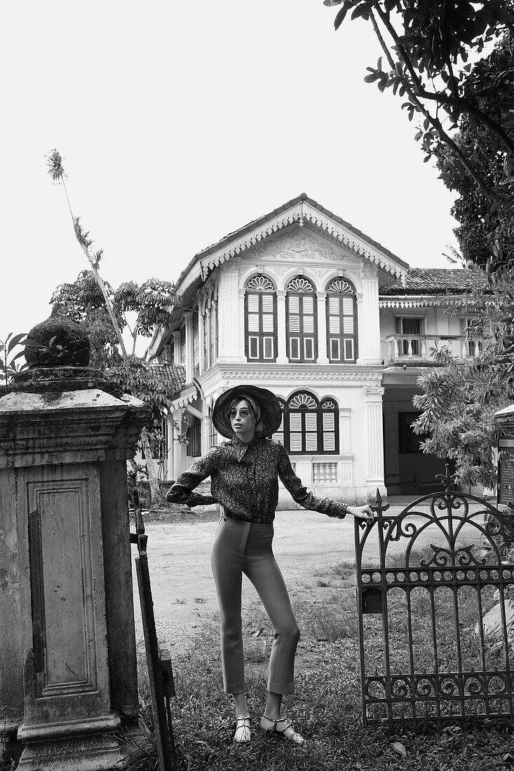 A young woman wearing a hat, trousers and a blouse standing in front of a house
