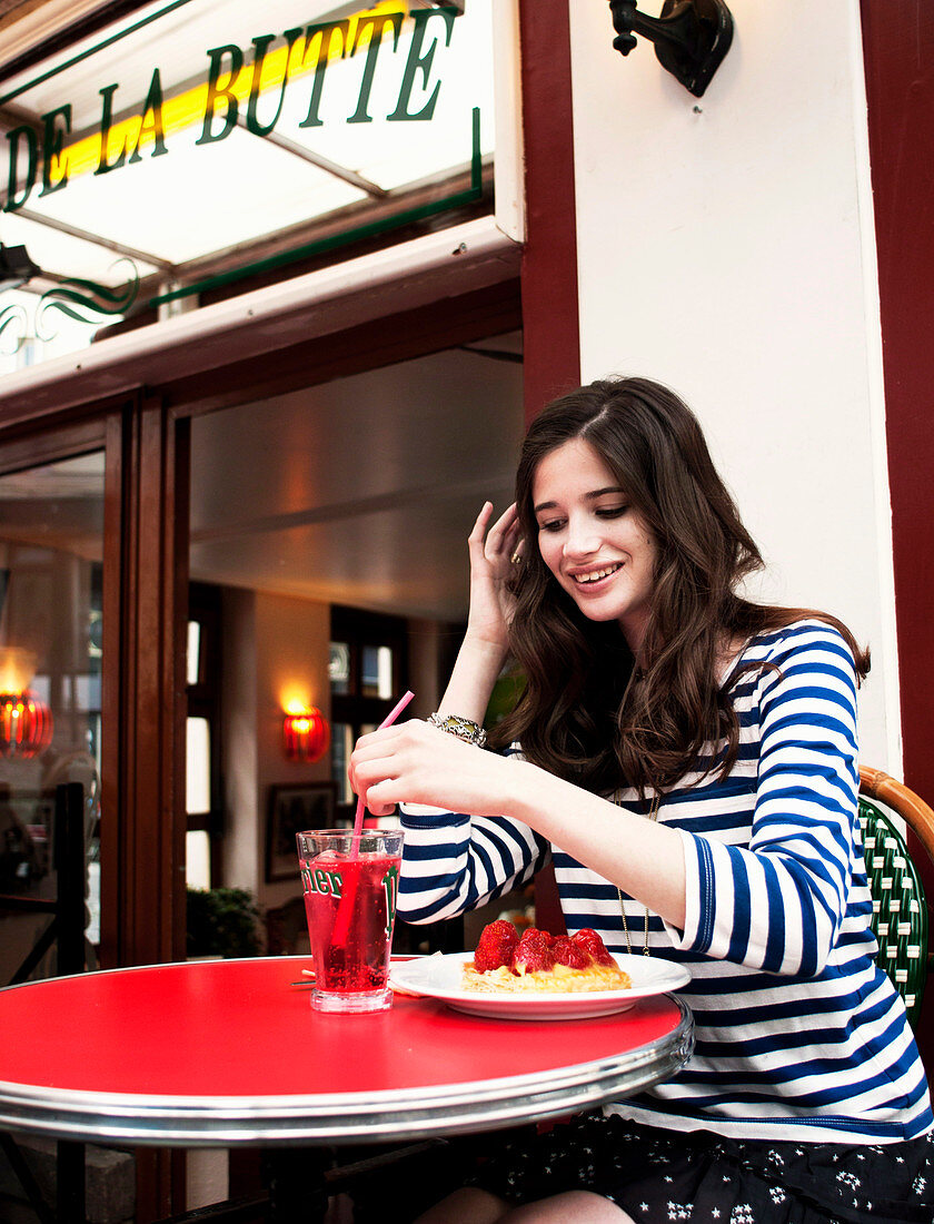Junge brünette Frau mit gestreiftem Shirt sitzt mit Kuchen und Getränk in einem Café