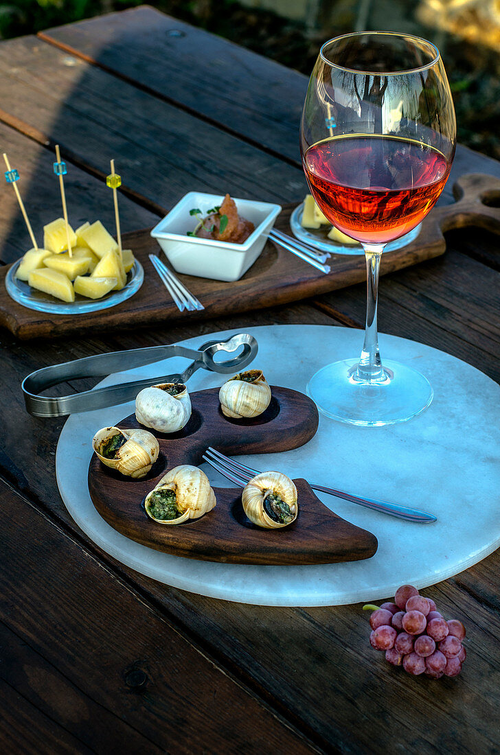 A glass of pink wine and appetizers - snails and cheese with chutney on a wooden table outdoors