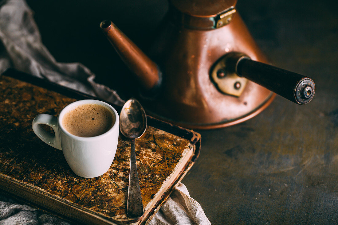 A cup of coffee on an antique book with an old-fashioned copper kettle in the background