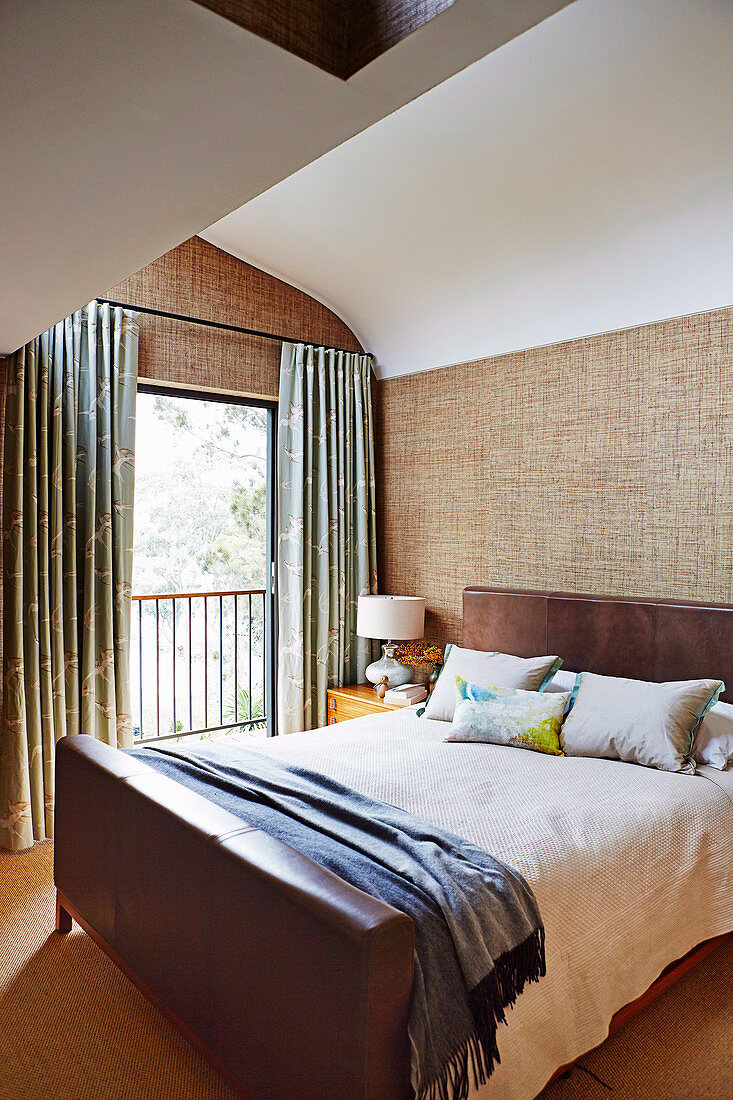 Double bed with leather headboard and footboard, textile wallpaper and French window in the bedroom