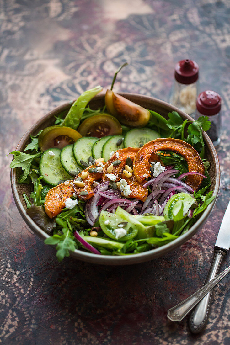 Salatschüssel mit Rucola, Gurke, Butternusskürbis, Tomate, roter Zwiebel, Pinienkernen und Ziegenkäse