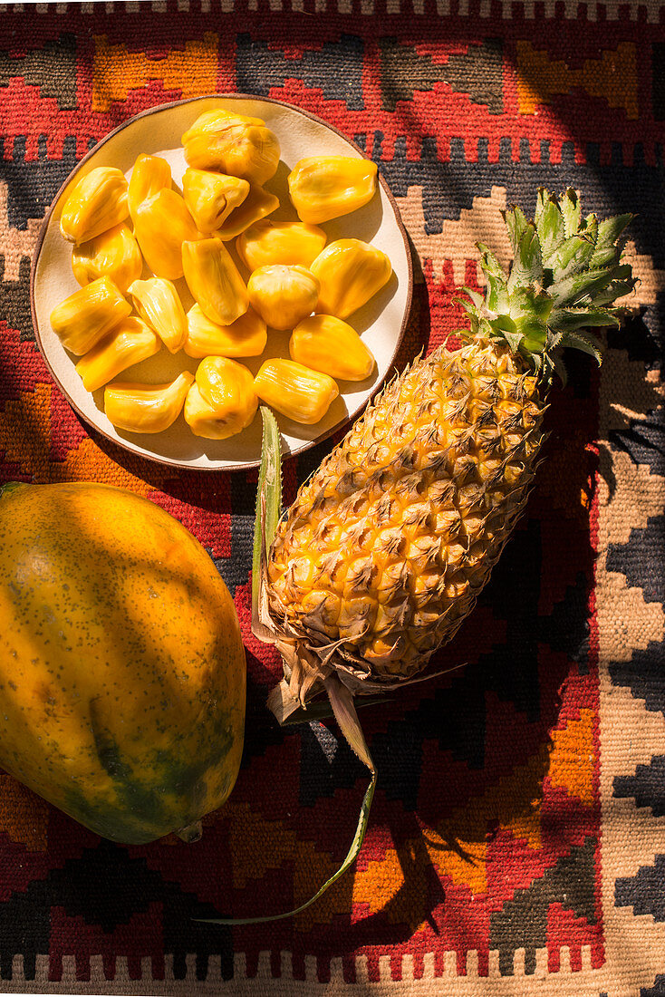 Jackfruit segments on a plate, whole pineapple and papaya on a colourful background