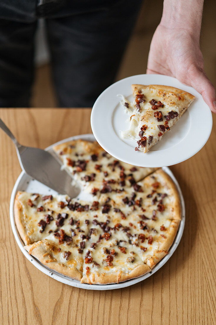 A person serving a slice of pizza with bacon and cheese on a plate