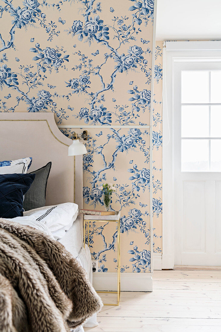 Double bed with headboard, wall-mounted lamp and bedside table in romantic bedroom with floral wallpaper