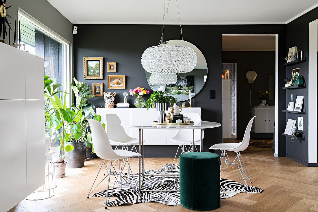 Filigraner Esstisch mit Klassikerstühlen auf Zebrafell, grüner Pouf und weiße Sideboards im Wohnraum mit dunklen Wänden