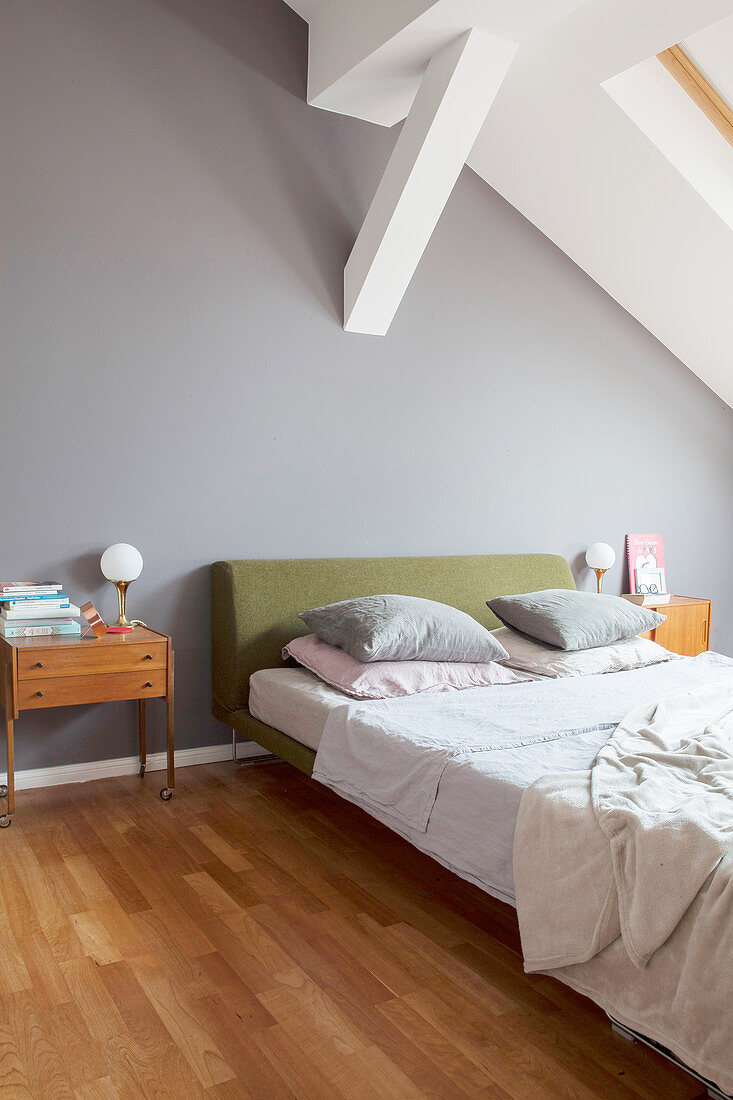 Double bed with green headboard and retro bedside table in bedroom with grey wall and white ceiling beams