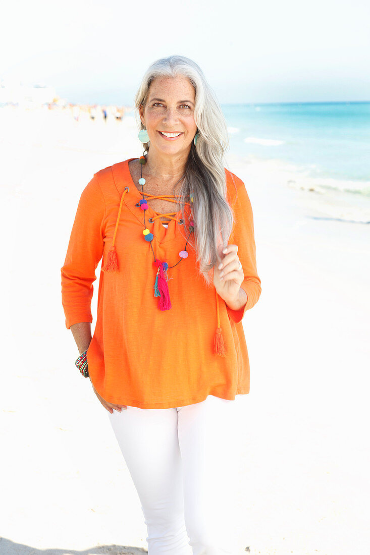 A mature woman with white hair on a beach wearing an orange tunic and white summer trousers