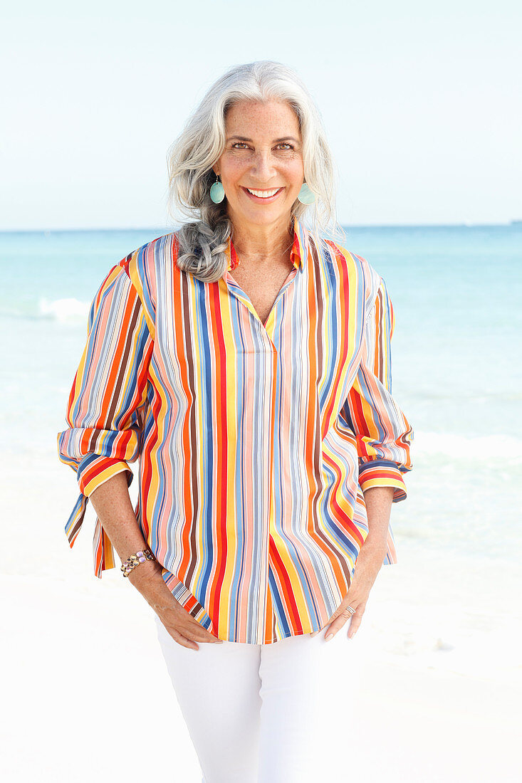 Reife Frau mit weißen Haaren in gestreiftem Hemd und weißer Sommerhose am Strand