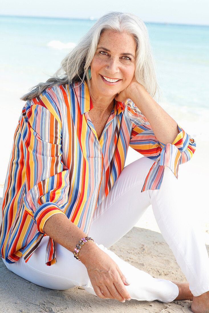 A mature woman with white hair on a beach wearing a striped shirt and white summer trousers