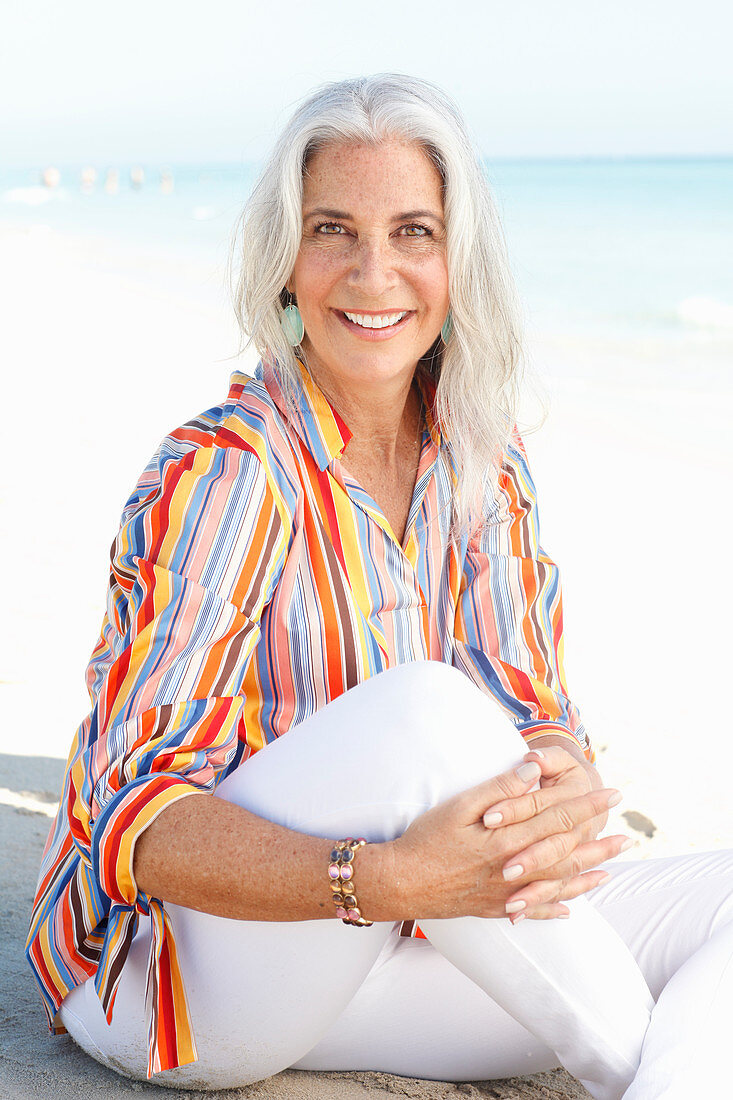 A mature woman with white hair on a beach wearing a striped shirt and white summer trousers