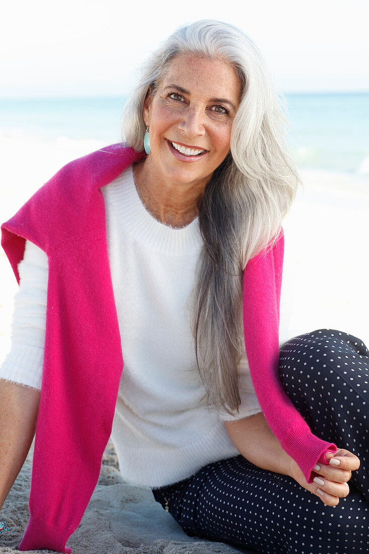 A mature woman with white hair on a beach wearing a white jumper and polka dot trousers with a pink jumper over her shoulders