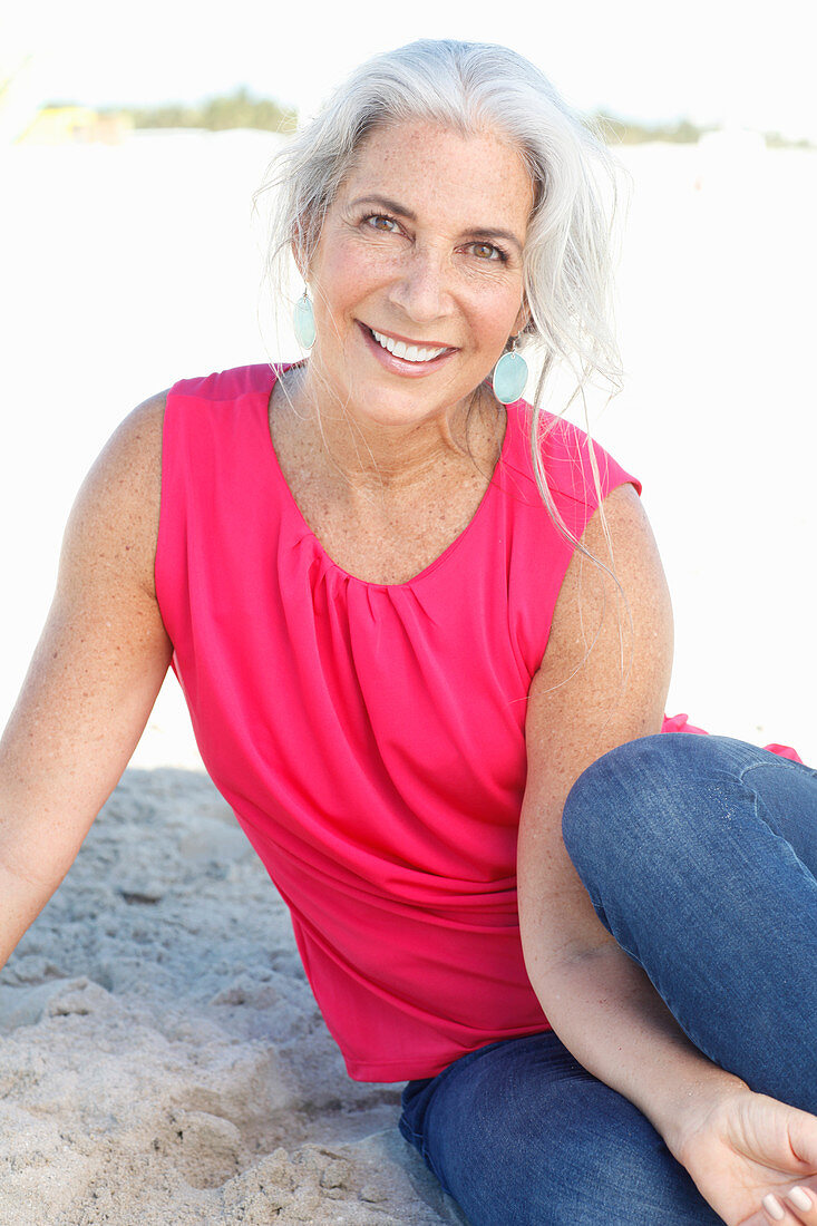 Reife Frau mit weißen Haaren in rosa Top und Bluejeans am Strand