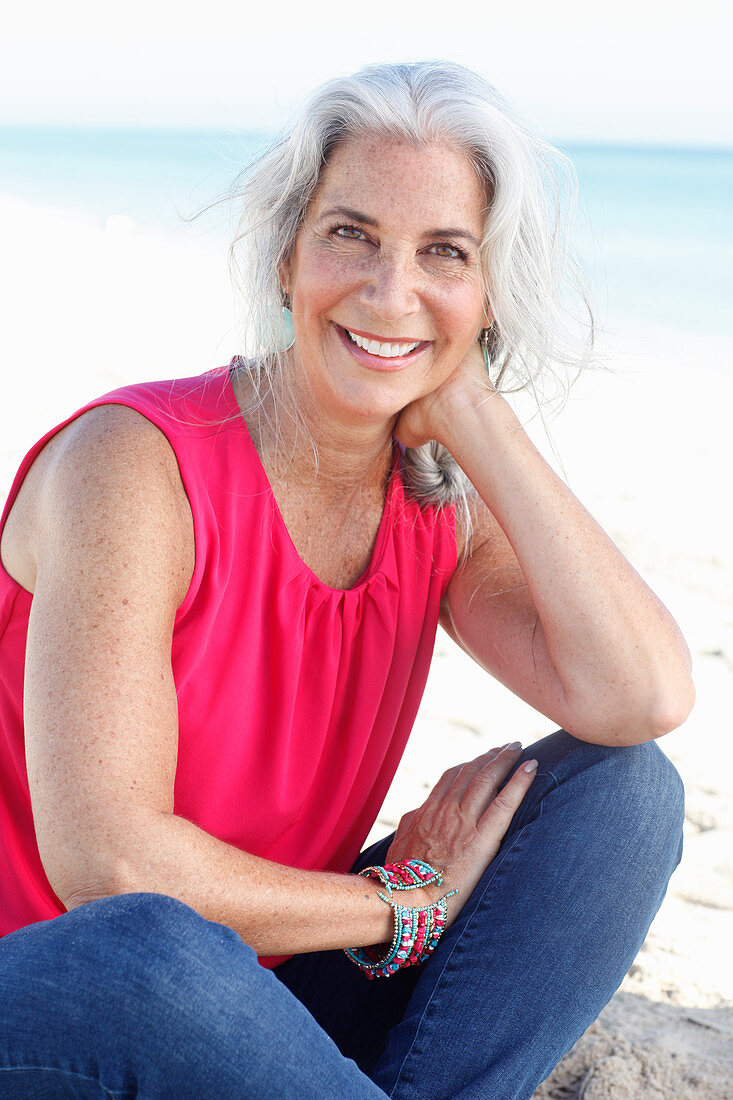 A mature woman with white hair on a beach wearing a pink top and blue jeans