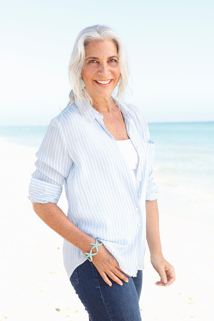 A mature woman with white hair on a beach wearing a striped blouse, a top and blue jeans