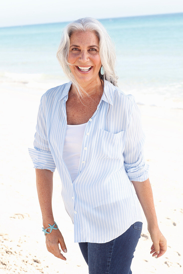 Reife Frau mit weißen Haaren in gestreifter Bluse, Top und Bluejeans am Strand