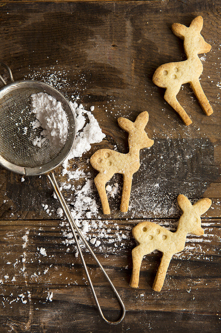 Three Festive Christmas Deer shaped cookie biscuits on a rustic board with icing sugar in a sifter