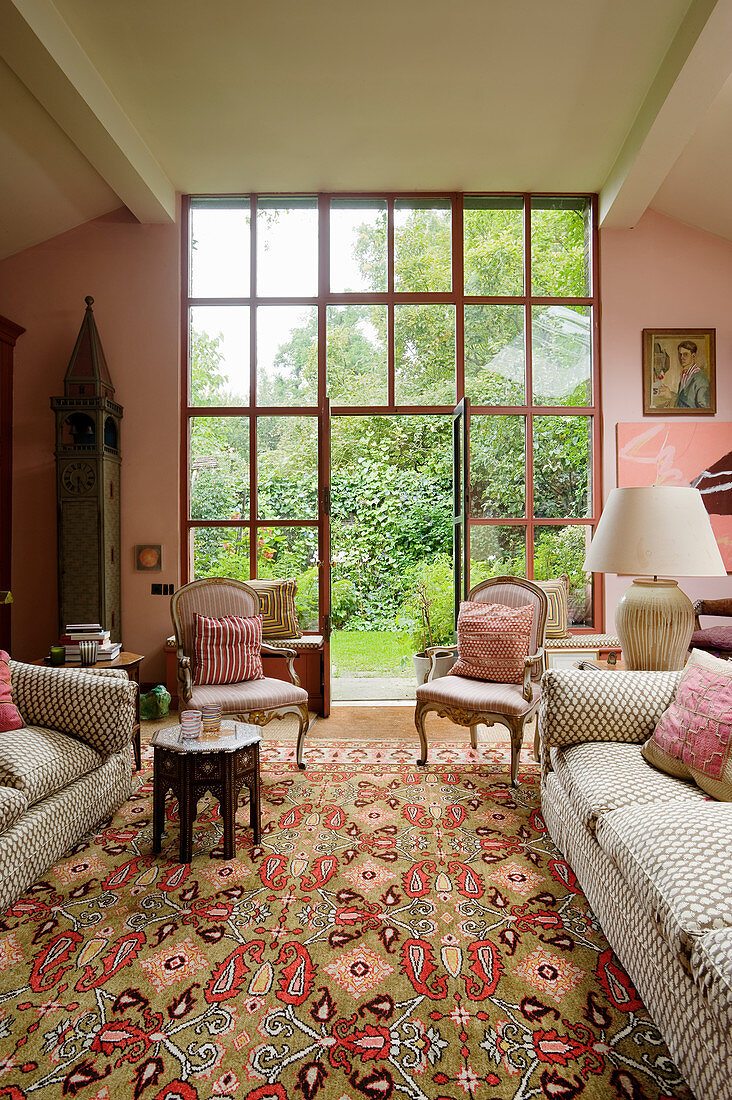 Couch and antique armchairs in front of floor-to-ceiling glass doors leading to garden in living room in shades of pink