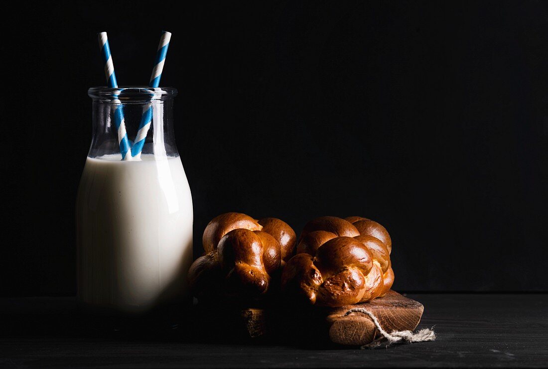 Eine Flasche Milch und Milchbrötchen auf rustikalem Holzbrett vor schwarzer Hintergrund