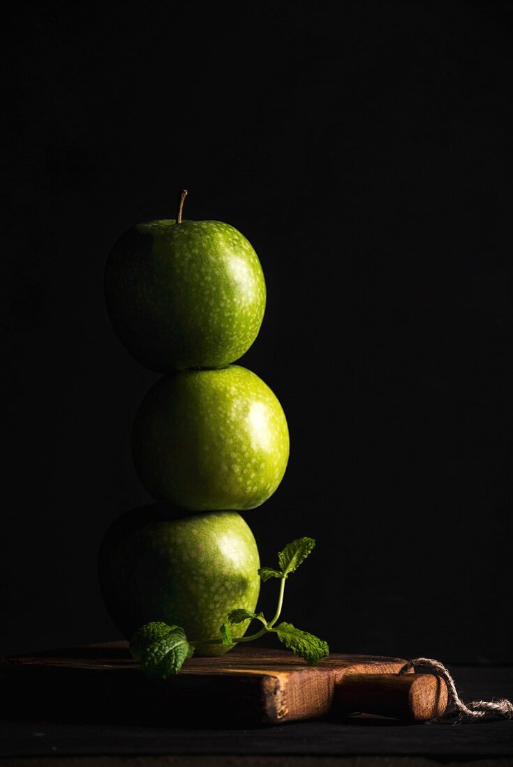 Gestapelte grüne Äpfel und Minzzweig vor schwarzem Hintergrund