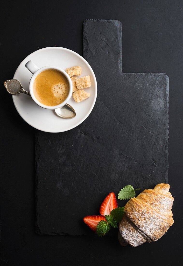 Eine Tasse Espresso und Croissant mit frischen Erdbeeren auf schwarzem Schieferbrett