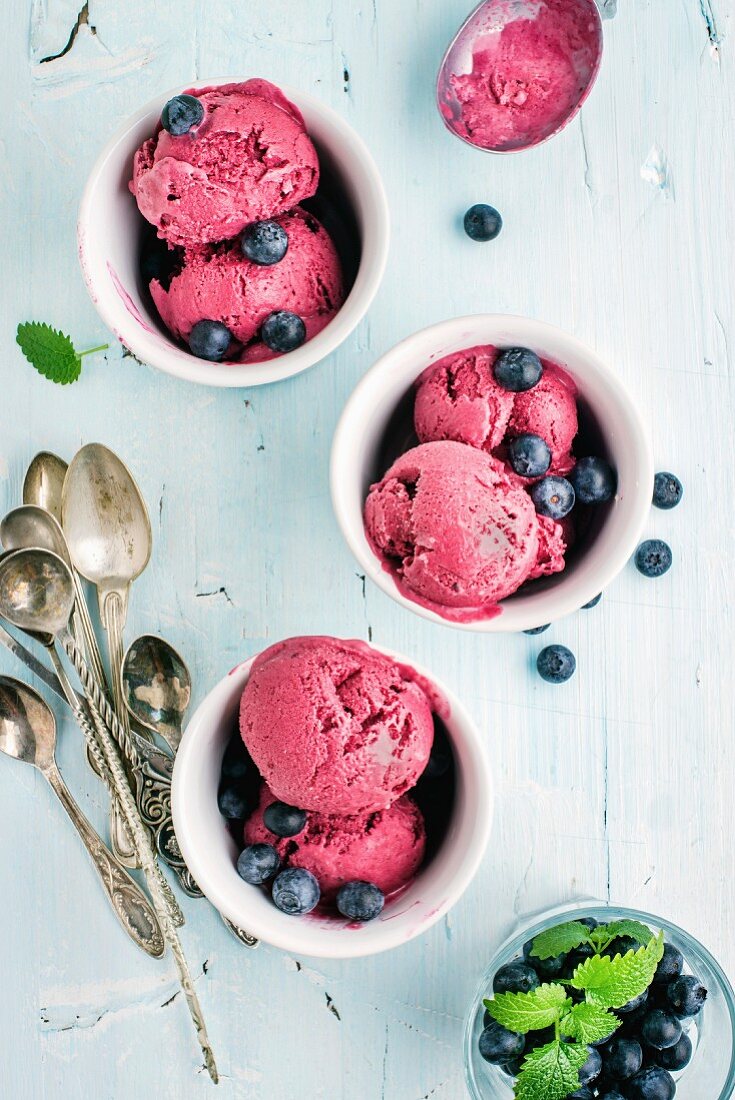 Homemade blueberry ice cream scoops with fresh berries and mint leaves in cups over light blue background