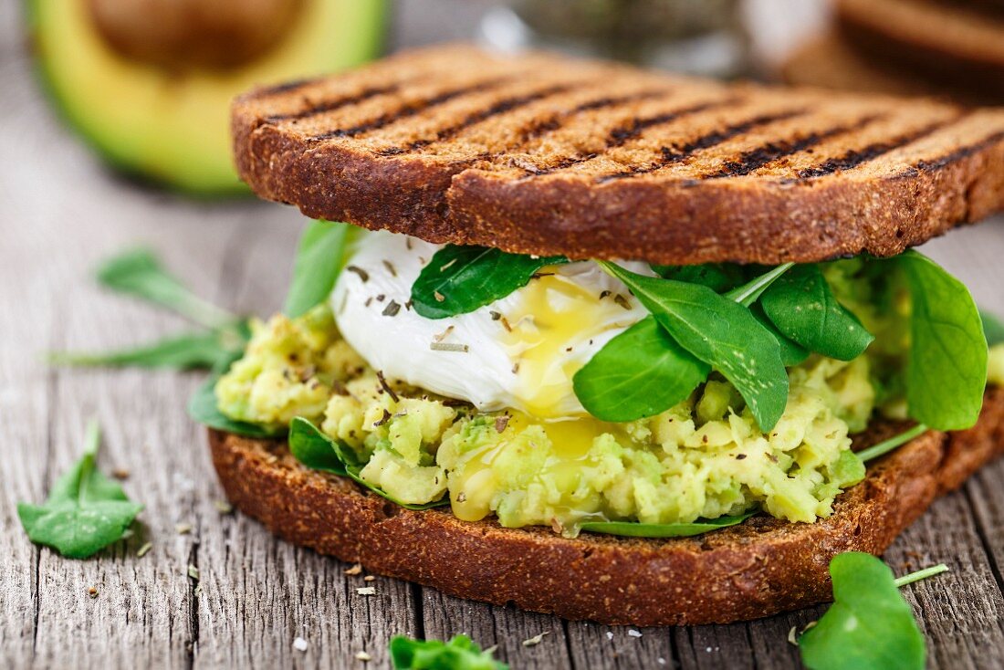 Grilled sandwich with avocado, poached egg and arugula on wooden table
