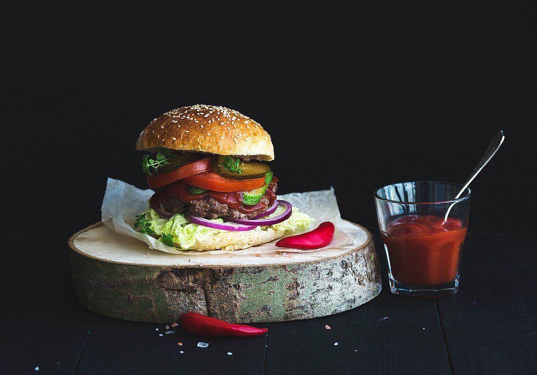 Selbst gemachte Burger auf Holzscheibe, daneben scharfe Tomatensauce im Glas