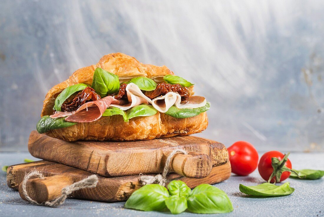 Croissant-Sandwich mit Parmaschinken, getrockneten Tomaten, frischem Spinat und Basilikum auf Holzbrett