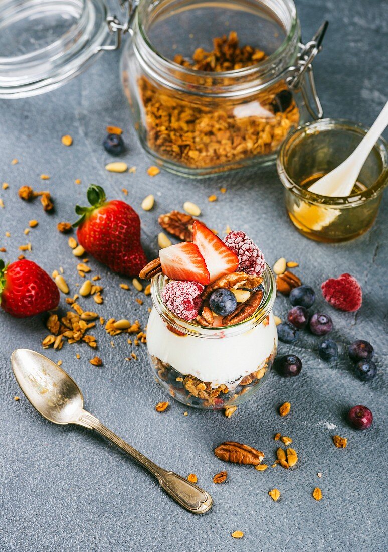 Yogurt oat granola with fresh berries, nuts, honey and mint leaves in glass jar on grey backdrop