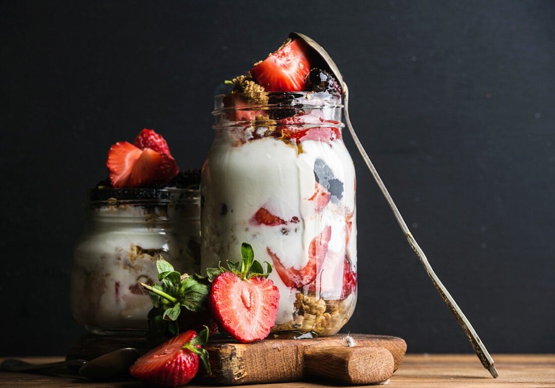 Yogurt oat granola with strawberries, mulberries, honey and mint leaves in tall glass jar on black backdrop