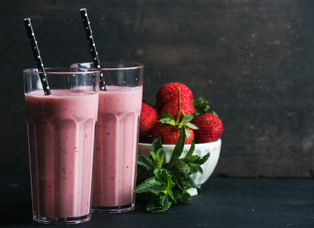Strawberry and mint smoothie in tall glasses, bawl of fresh berries on dark rustic wood background