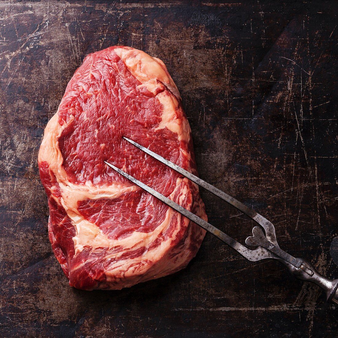 Rohes Ribeye Steak und Fleischgabel auf dunklem Hintergrund