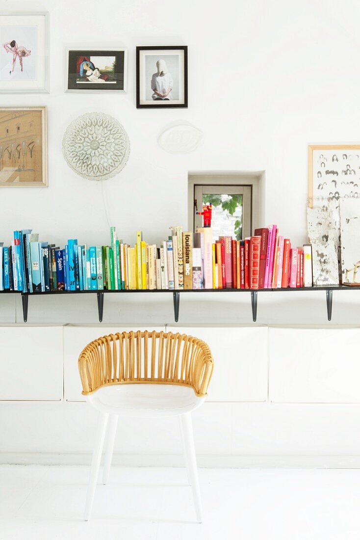 Weißer Korbstuhl vor farblich sortierten Büchern auf Wandboard