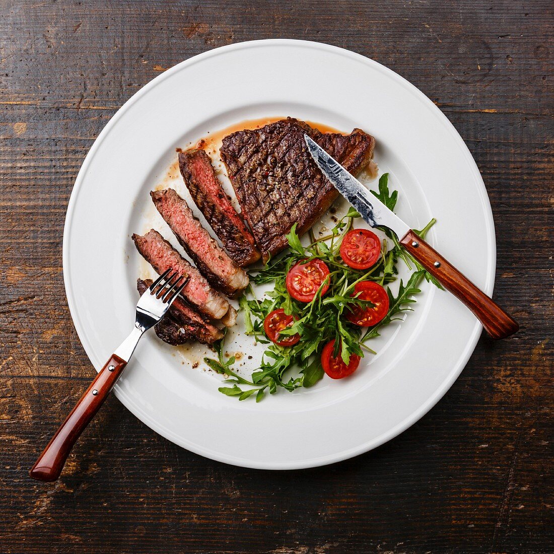 Sliced medium rare grilled Beef steak Striploin and salad with tomatoes and arugula on white plate