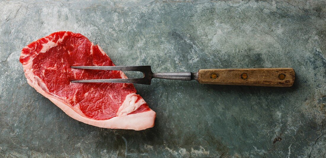 Raw fresh meat steak Striploin and meat fork on gray stone slate background