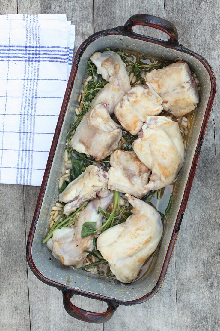 Kaninchen im Bräter mit Schalotten, Knoblauch, Kräuterzweigen und Pinienkernen