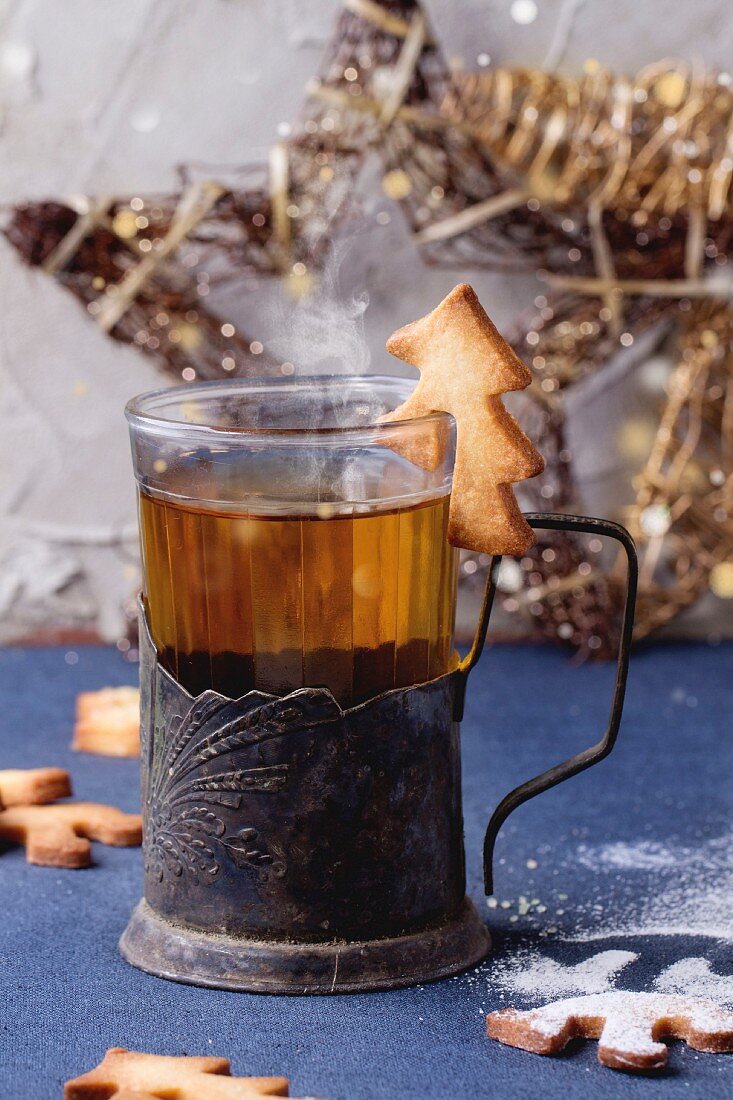 Shortbread Christmas cookies for cups in sugar powder, glass of hot tea in cup holder on table with blue tablecloth