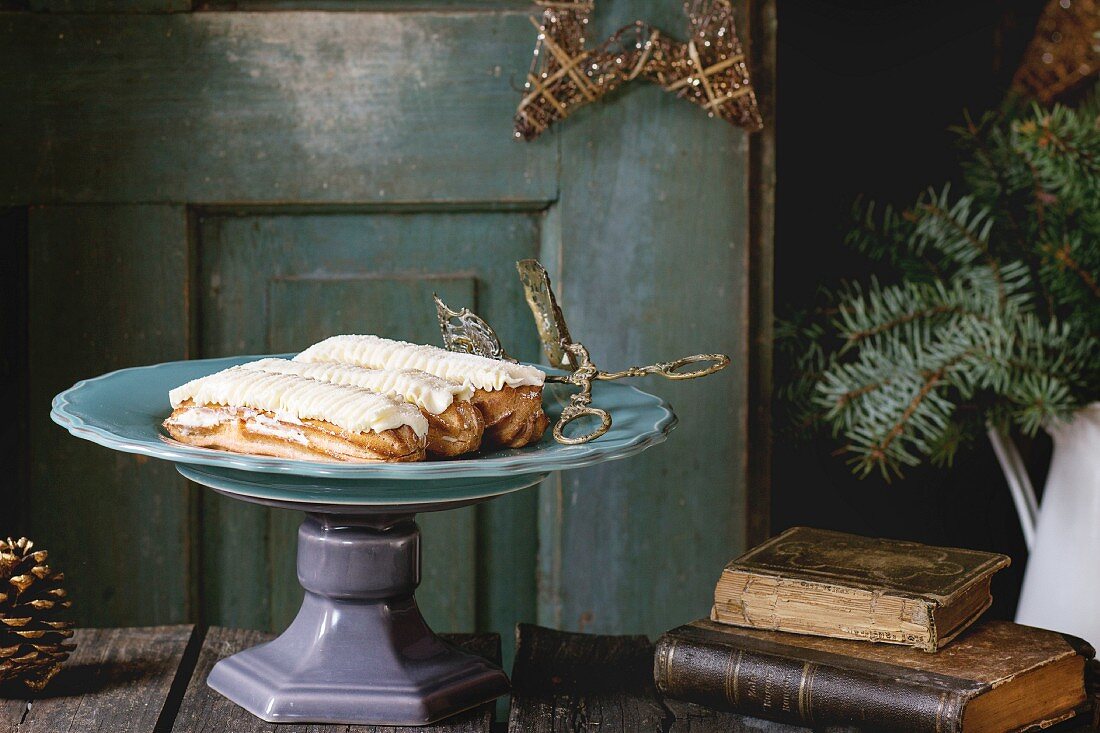 Old wooden Christmas table with big plate of Butter cream Eclairs, decorating by Christmas stars, tree and vintage books