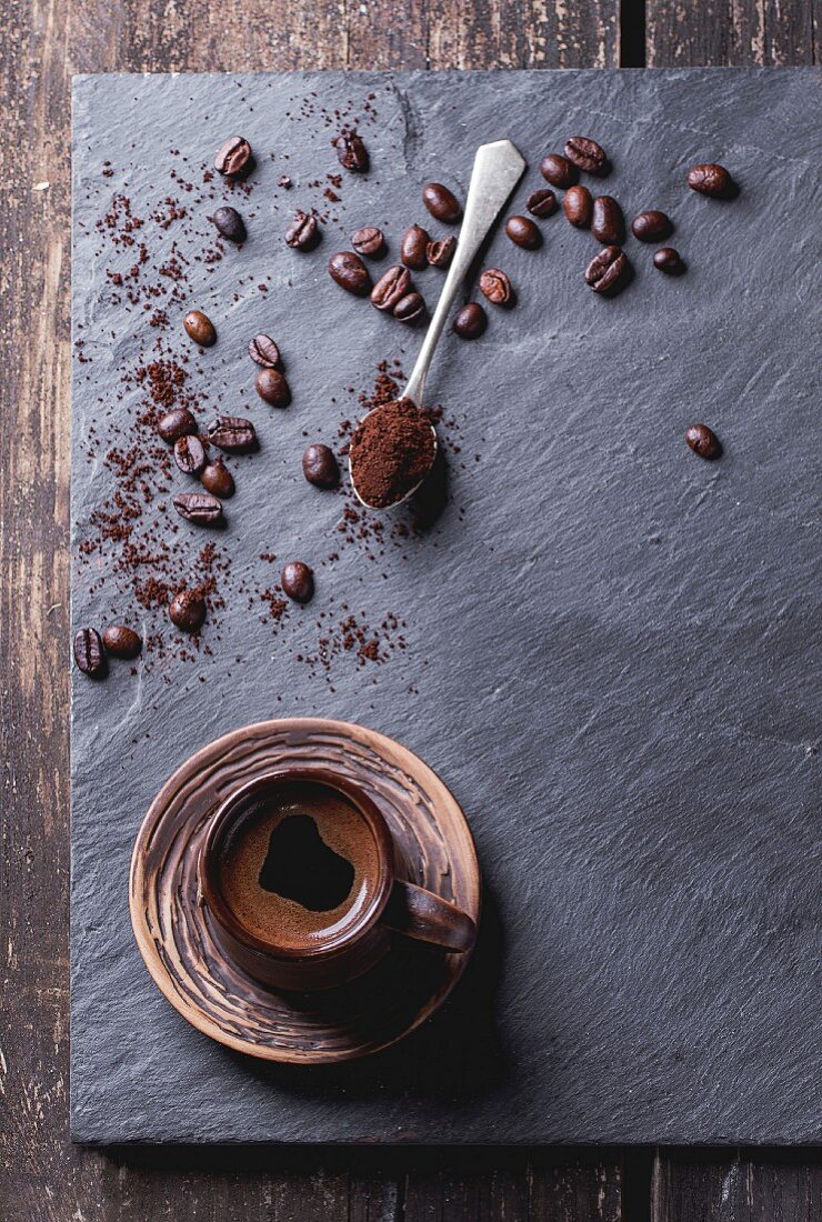 Brown ceramic cup of fresh coffee, tea spoon with ground coffee and coffee beans over black slate as background