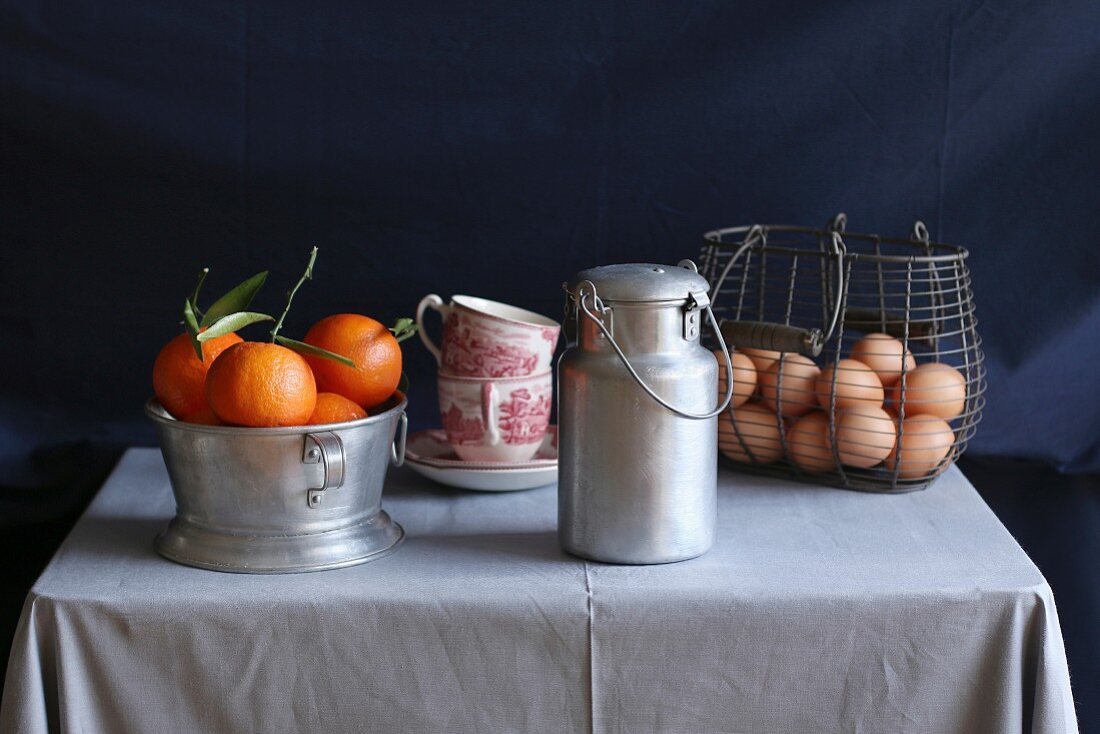An arrangement of clementines, vintage coffee cups, an aluminium milk jug and eggs