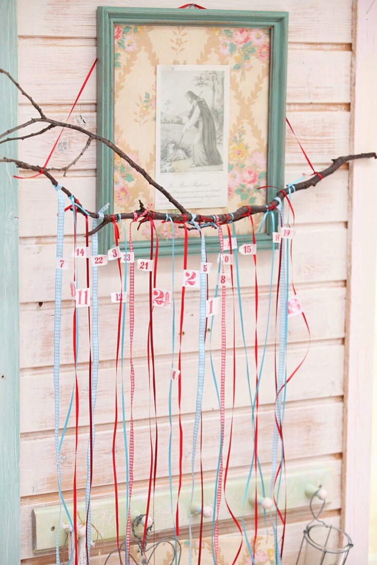 DIY-Adventskalender mit nummerierten Geschenkbändern an Ast vor Heiligenbild aufgehängt