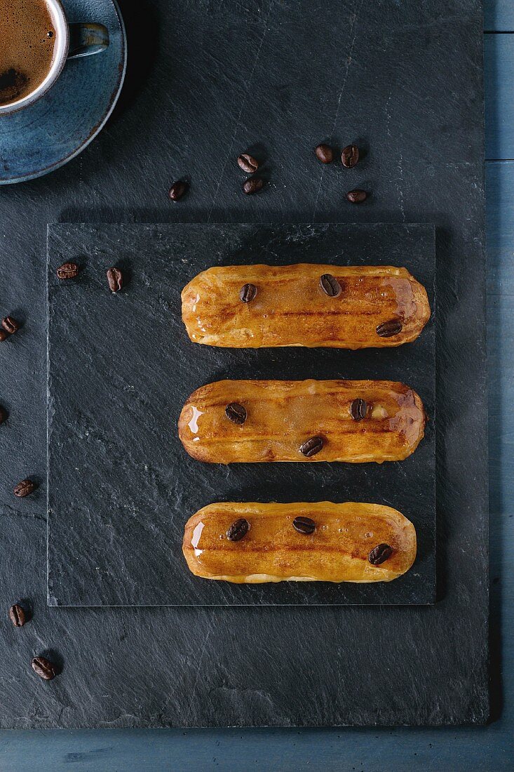 Drei Kaffee-Eclairs mit Tasse Kaffee und Kaffeebohnen auf schwarzer Schiefertafel