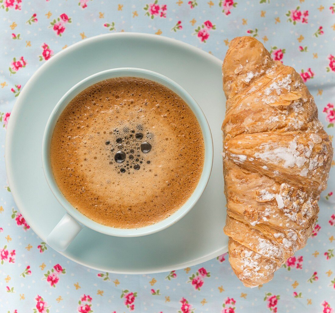 Frothy coffee in a blue cup and saucer on a floral table cloth, with croissant pastry