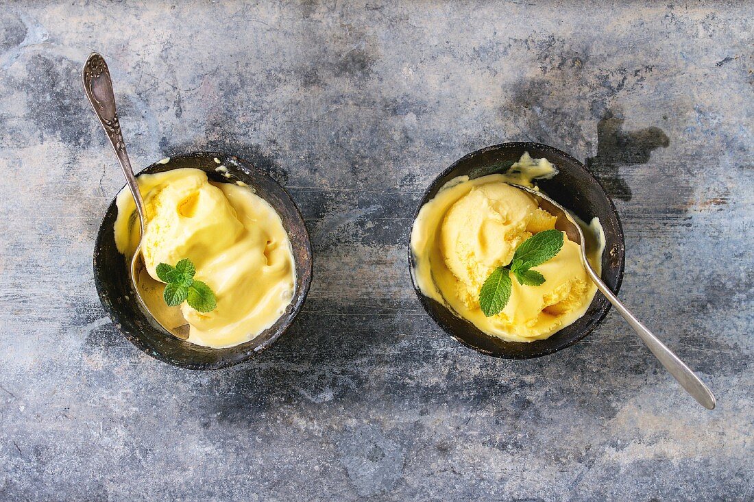 Homemade mango ice cream with fresh mint in vintage iron bowls over gray metal textured background