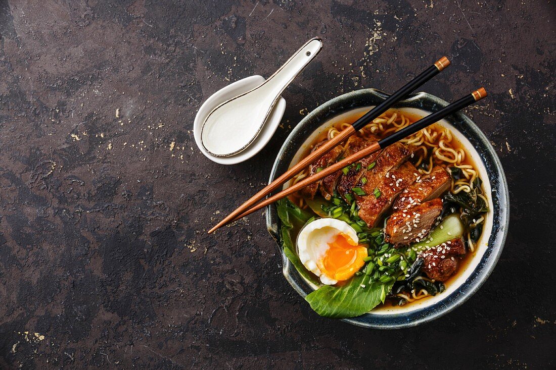 Duck noodles with egg and pak choi cabbage in bowl on dark black stone texture background